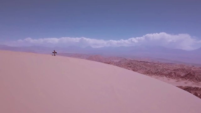 Man climbing upholding a sandboard over great Sand Dunes of the Dead Valley, Atacama desert. Aerial view, 4k