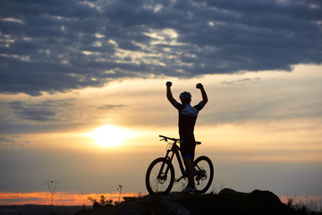 Man in sports clothes and a helmet stands near cycle with his hands up on a rock on top of a mountain with a beautiful landscape of the sky at sunset and hills in the distance. Back view