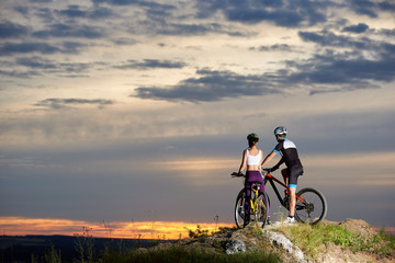 Fototapeta na wymiar Back view of a couple riding a mountain bike on top enjoying the sunset and a beautiful evening in the mountains. Young people dressed in sporting clothes and helmets