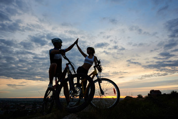 Obraz premium Athletic man and sttractive woman in helmets highing five and looking at each other after cycling. Sporty couple of cyclists sitting on bikes and resting in twilight. Evenening sky background.