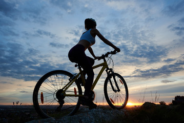 Fototapeta na wymiar Back view of young woman cycling mountain bicycle. Silhouette of female cyclist in helmet enjoys sunset on a top of mountain under fairytale sky with clouds and bright sun in the evening