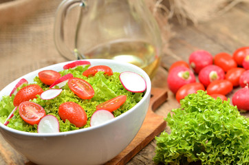 Close-up of green salad Lollo Biondo with tomatoes and radishes in a white bowl on wooden table, a pitcher of oil in the background
