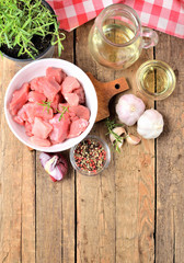VIew from above of raw pork meal in a bowl, garlic, onion, rosemary in a pot, pepper, jug with oil and checkered red tablecloth in the background - vertical photo