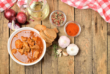 View from above of pork goulash with pieces of meat in a bowl, garlic, pepper, onion, jug with oil and red checkered tablecloth in the background