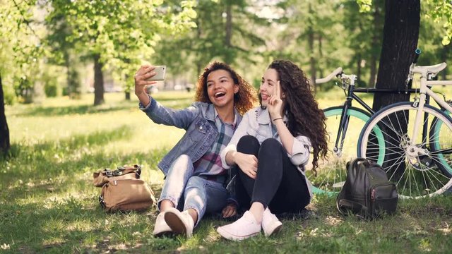 Pretty girls friends are taking selfie in park sitting on lawn with bicycles in background. Mixed race friendship, modern technology and cheerful people concept.