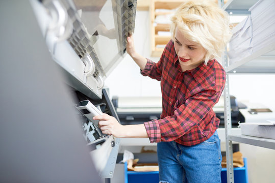 Beautiful young woman in casual outfit inspecting plotter while working in printing office. 