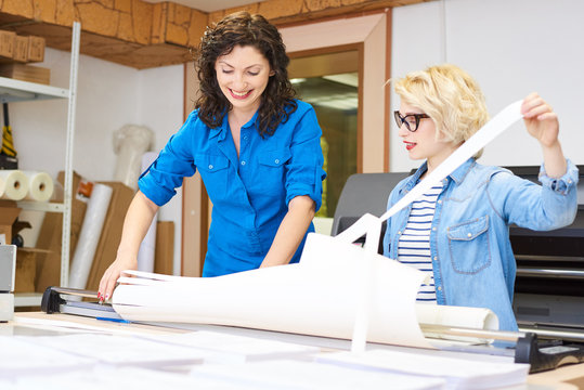 Two pretty women smiling and tearing plotter paper on stripes while working in printing office. 