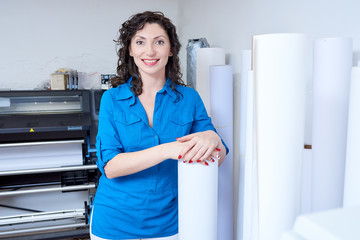 Beautiful woman in blue shirt smiling and looking at camera while leaning on paper roll in printing office. 
