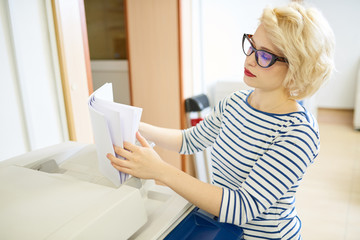 Lovely young woman in glasses standing near plotter and sorting stack of paper while working in...