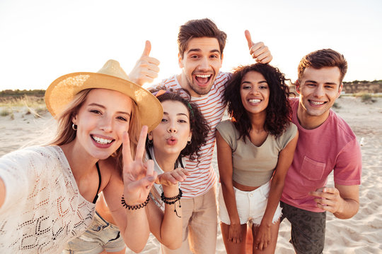 Photo of happy young multiracial people in summer clothes taking selfie, while standing at the beach during sunset