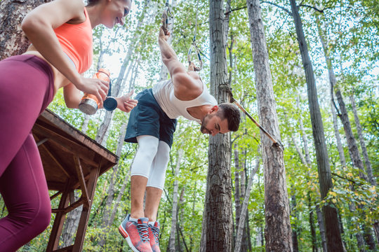 Woman and man doing athletic training in the woods at rings, she is cheering him 