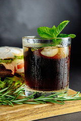 Glass of cola with ice, mint and  rosemary on black background  big sandwich, street food