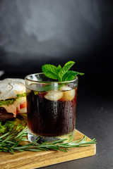 Glass of cola with ice, mint and  rosemary on black background  big sandwich, street food