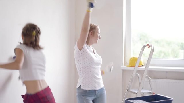 Mother and daughter painting wall in apartment room. Beautiful family doing repairs in new flat. Funny woman and teen girl having good mood - making decorate room and dancing.