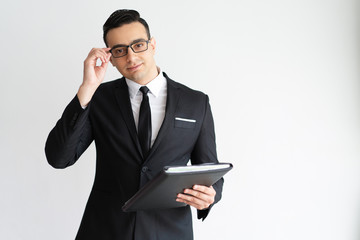 Serious handsome young businessman adjusting glasses and holding folder. Content successful business expert in formal suit looking at camera. White collar concept