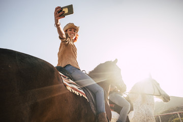 couple riding horses take a selfie with modern technology smartphone. cowboy lifestyle and smile woman. sunset time and backlight for outdoor leisure activity for friends