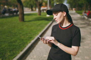 Young athletic beautiful brunette girl in black uniform and cap using mobile phone during training,...
