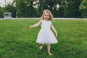 Smiling little cute child baby girl in light dress walking and running, have fun on green grass lawn in park. Mother, little kid daughter. Mother's Day, love family, parenthood, childhood concept.