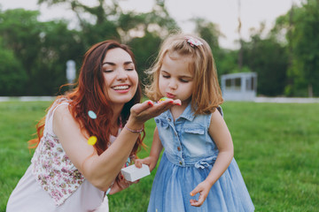 Fototapeta premium Woman in light dress and little cute child baby girl play and blowing on colorful paper confetti in green park. Mother, little kid daughter. Mother's Day, love family, parenthood, childhood concept.