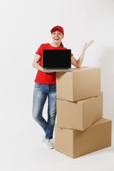 Full length of delivery woman in red cap, t-shirt isolated on white background. Female courier near empty cardboard boxes, laptop pc computer, blank black empty screen. Receiving package. Copy space.