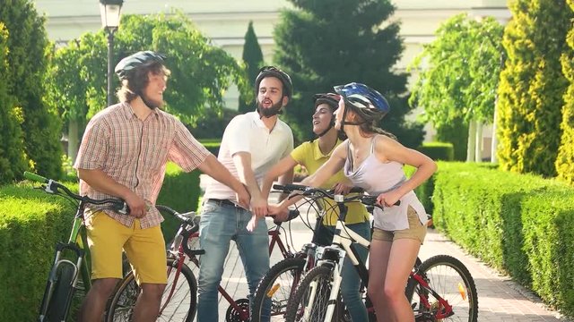 Four happy friends stacking hands outdoors. Young cheerful cyclists resting in park, slow motion. Students on summer holidays with bikes.
