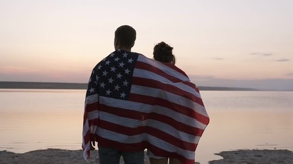 Close-up of beautiful young couple looking forward and dreaming. Embraced lovers thinking about their future life while standing by the sea in the calm evening, covered with american flag - Powered by Adobe