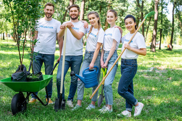 young volunteers posing in park with watering can, shovel, rake and wheelbarrow with new trees