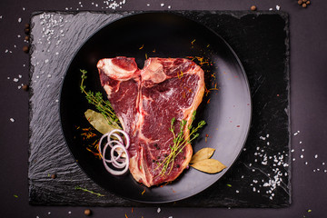 Raw meat, a beef steak, pork or veal, lamb, lies on a black ceramic plate, on a black slate. Next to the meat are spices and vegetables. Top view. Copy space