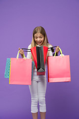Child look in shopping bags on violet background. Little shopaholic smile surprised with paper bags. Childhood. Shopping and sale. Shopper girl with paperbags. What a surprise. Big sale and shopping