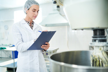 Confectionery factory employee standing in white coat near operating machinery and making notes. 