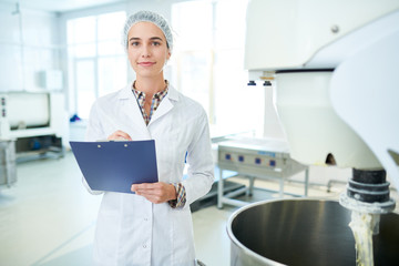 Confectionery factory employee standing in white coat near operating machinery holding clipboard...