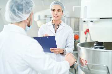 Confectionery factory employee standing near machinery in white coat making notes and speaking with...