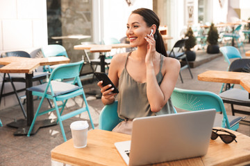 Image of modern brunette woman sitting in city cafe or restaurant outside in summer, and using cell phone with bluetooth wireless earphone