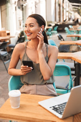 Image of adorable brunette woman 20s sitting in city cafe or restaurant outside in summer, and touching bluetooth wireless earpbud with cell phone in hand