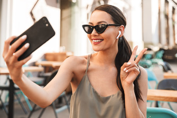 Beautiful brunette woman 20s sitting in city cafe or restaurant outside in summer, and taking selfie photo on smartphone with bluetooth wireless earpbud in ear