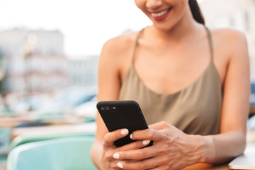 Cropped image of smiling brunette woman looking at mobile phone holding in hands, while sitting in city cafe or restaurant outside in summer