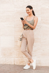Image of pleased brunette woman wearing casual summer clothing looking at cell phone holding in hands, while standing against beige wall outside