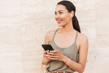 Portrait of smiling brunette woman wearing casual summer clothing and bluetooth earpod looking...