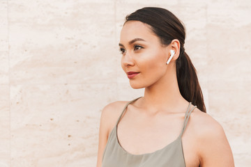 Portrait of beautiful brunette woman wearing casual summer clothing and bluetooth earpod looking aside, while standing against beige wall outdoor