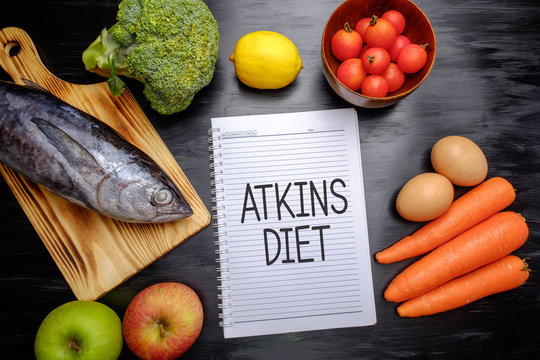 Atkins Diet on chalkboard, health conceptual. Healthy fresh food fish, lemon, tomatoes, apple, carrot and broccoli.