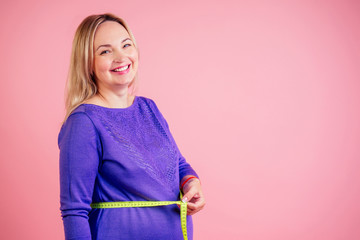 Beautiful blonde smiley pregnant woman in a dress with red satin bow on the belly measures baby bump with measuring tape in the studio on a pink background copyspace