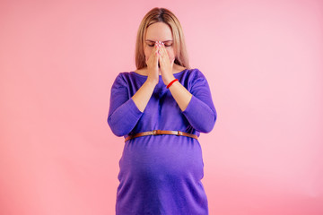 Beautiful blonde pregnant sad woman in a violet dress with big baby bump offended and upset headache in the studio on a pink background