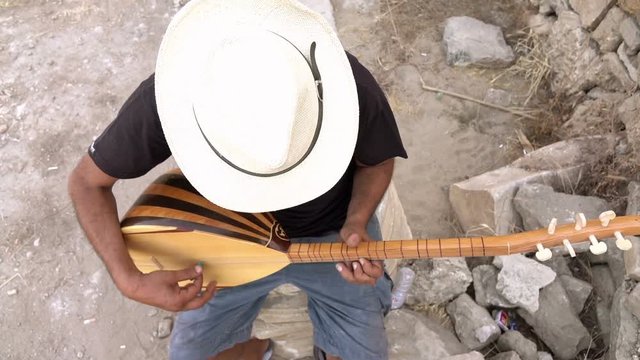 A street musician plays a Turkish instrument. A man in a white hat plays a game of baglama.