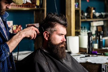 Barber with hair clipper works on hairstyle for man with beard, barbershop background. Barber...