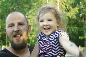 A father with a beard is holding a daughter in his arms, they are fooling around, smiling. Joyful and happy family portrait