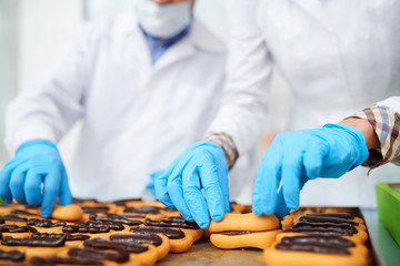 Crop close-up confectionery factory workers in protective gloves preparing pastry with chocolate...