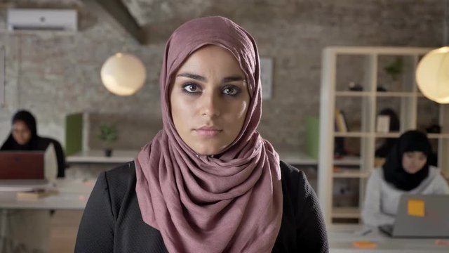 Portrait of young muslim women in hijab looking in camera, serious and concerned, three pretty womens working on laptop in modern office