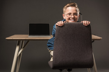 happy little boy sitting on chair and looking at camera near table with lamp and laptop on grey background