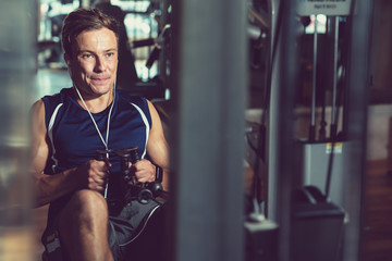 Fototapeta na wymiar Concentrated young sportsman using rowing machine and listening to music in headphones while having intensive training at gym, portrait shot