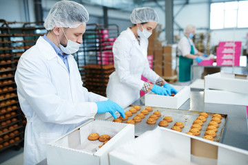 Confectionery factory employees in white coats collecting freshly baked pastry from tray and...